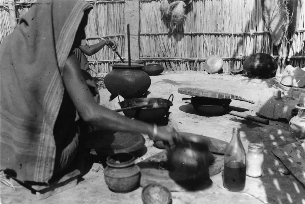 Black-and-white photograph of women cooking in an outdoor courtyard kitchen in Dhaka, with clay pots and a grinding stone